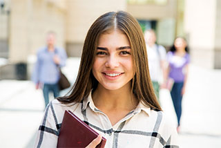 Smiling young woman holding a notebook outdoors on a college campus.