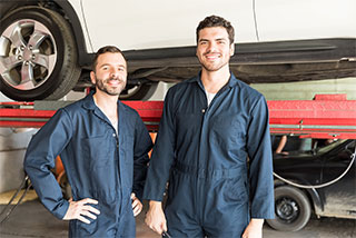 Two smiling auto mechanics in blue coveralls standing in a garage with a car on a lift.