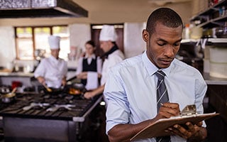Restaurant manager taking inventory in commercial kitchen with chefs cooking in background