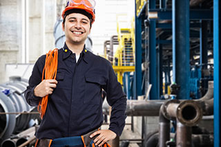 Smiling electrician wearing a hard hat and work uniform, holding electrical cables in an industrial setting.