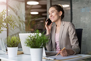 Smiling businesswoman in a modern office, talking on the phone while working on a laptop and taking notes.