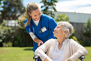 Smiling health care aide in blue scrubs helping a happy senior woman in a wheelchair outdoors.