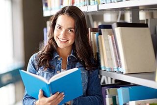 Smiling young woman in a denim jacket reading a book in a library.