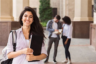 Smiling female student with a backpack and books standing outside a campus.