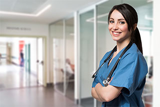 Smiling nurse in blue scrubs with her arms crossed standing in a hospital hallway.