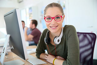 Female student in red glasses with headphones around her neck at a desk.